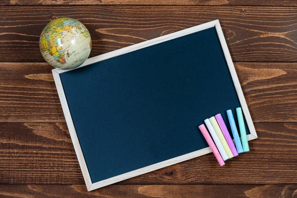 A blackboard with space for text, a globe and a set of colored crayons on a wooden background. Start of school year and sale of school supplies. Home education and learnin after pandemic. Flatlay. — Stock Photo, Image