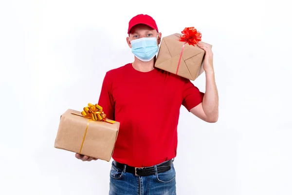 Bezpečná bezkontaktní dálková dodávka rekreačních dárků během pandemie koronaviru. Kurýr v červené uniformě a ochranné lékařské masce obsahuje dvě krásné dárkové krabice s luky. — Stock fotografie