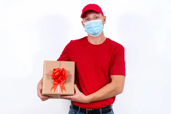 Safe delivery of gifts for holidays. A courier in red uniform and protective medical mask holds box with a bow. Contactless remote gift orders in quarantine during the coronavirus pandemic. — Stock Photo, Image