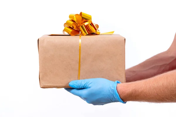 Entrega remota segura y sin contacto de regalos navideños durante la pandemia del coronavirus. De cerca. La mano de mensajero en guantes médicos protectores sostiene una hermosa caja de regalo con un lazo sobre un fondo blanco. — Foto de Stock