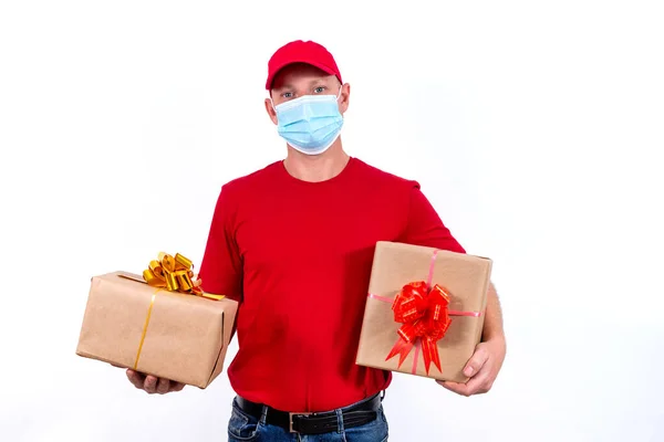Livraison à distance sans contact de cadeaux pendant la pandémie de coronavirus. Un messager dans un uniforme rouge et un masque médical de protection détient deux belles boîtes-cadeaux avec des arcs. — Photo