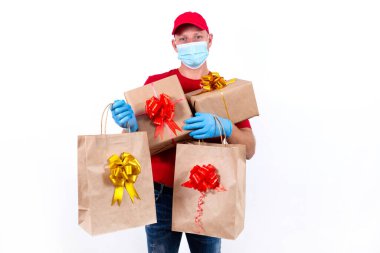 Coronavirus salgını sırasında uzak mesafeden tatil hediyelerini güvenli bir şekilde teslim etmek. Kırmızı üniformalı, koruyucu tıbbi maskeli ve eldivenli bir kurye büyük bir sipariş, bir sürü hediye kutusu ve fiyonklu çanta taşır..