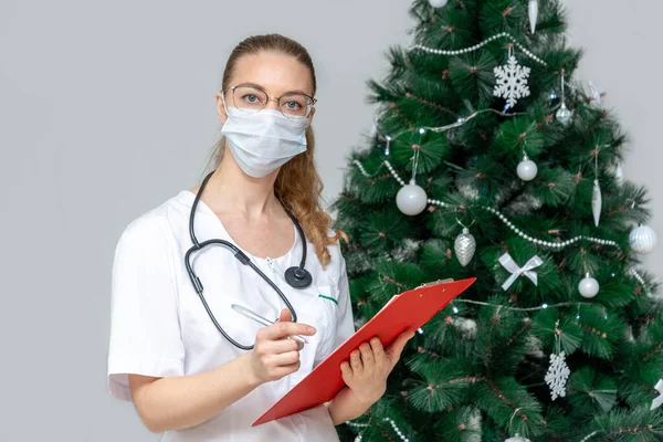 A female doctor in a protective medical mask holds a clipboard in background of a Christmas tree. Make an appointment at hospital in the new year and Christmas. Online reception and patient care.
