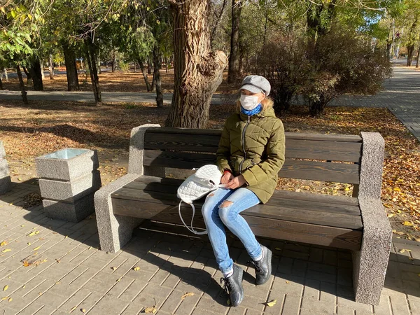 Mulher caucasiana em máscara protetora senta-se em um banco na rua em um parque de outono vazio. Estilo de vida e comportamento seguro durante a pandemia do coronavírus. Nova realidade normal e moderna. medidas de segurança — Fotografia de Stock