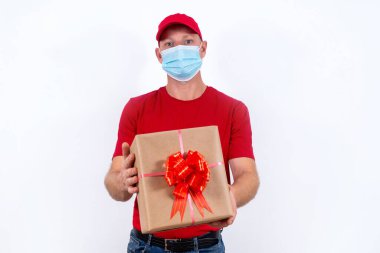 Tatil için güvenli bir hediye teslimatı. Kırmızı üniformalı ve koruyucu tıbbi maskeli bir kuryenin elinde fiyonklu bir kutu var. Koronavirüs salgını sırasında karantinada uzaktan kumandalı hediyeler..