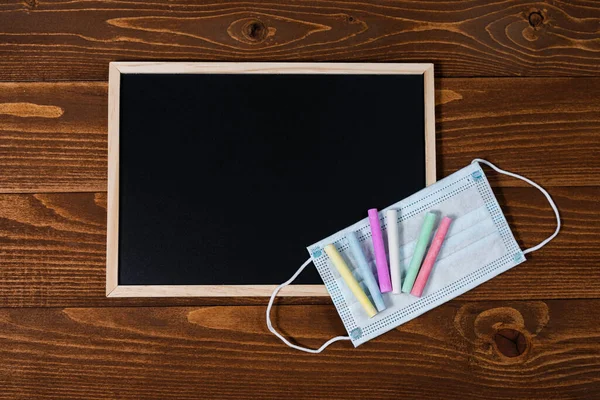 A blackboard with space for text, set of colored crayons and a protective mask on a wooden background. Start of school year and sale of school supplies. Home education and learnin after pandemic.