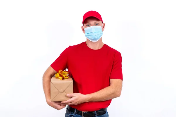 Safe delivery of gifts for holidays. A courier in red uniform and protective medical mask holds box with a bow. Contactless remote gift orders in quarantine during the coronavirus pandemic. — Stock Photo, Image