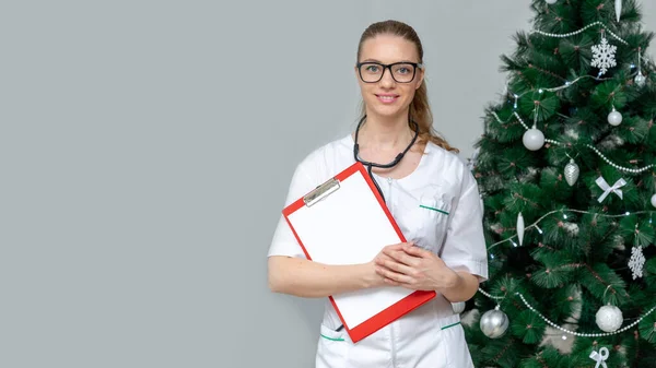 Banner. A female doctor holds a clipboard in background of a Christmas tree. Make an appointment at hospital in new year and Christmas. Online reception and patient care. Copy space. Space for text.