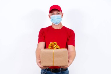Tatil için güvenli bir hediye teslimatı. Kırmızı üniformalı ve koruyucu tıbbi maskeli bir kuryenin elinde fiyonklu bir kutu var. Koronavirüs salgını sırasında karantinada uzaktan kumandalı hediyeler.