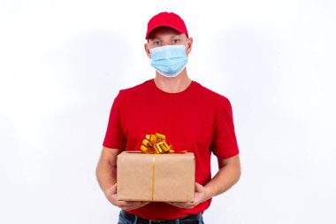 Tatil için güvenli bir hediye teslimatı. Kırmızı üniformalı ve koruyucu tıbbi maskeli bir kuryenin elinde fiyonklu bir kutu var. Koronavirüs salgını sırasında karantinada uzaktan kumandalı hediyeler.