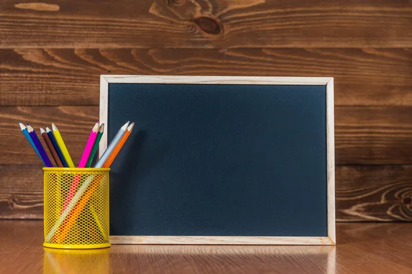 A blackboard with space for text, a set of colored pencils with a glass on a wooden background. Concept start of the school year and sale of school supplies. Home education and learning. — Stock Photo, Image