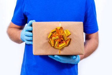 Coronavirüs salgını sırasında uzak mesafeden tatil hediyelerini güvenli bir şekilde teslim etmek. Kapatın. Koruyucu tıp eldivenleri içindeki kurye elinde beyaz arka planda fiyonklu güzel bir hediye kutusu var..