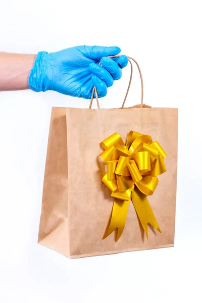 코로나 바이러스가 유행하는 동안 무선으로 선물을 멀리서 안전하게 배달하는 거죠. 닫아. 보호가 되는 의료용 장갑을 든 배달부가 흰 바탕에 활을 단 종이 선물 주머니를 들고 있다. — 스톡 사진