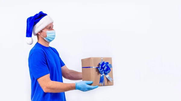 Koerier in blauw uniform, beschermend medisch masker, handschoenen en Kerstman hoed houdt geschenkdoos. Gelukkig nieuwjaar en vrolijk kerstfeest. Veilige contactloze levering op afstand van vakantiegeschenken tijdens het coronavirus. — Stockfoto