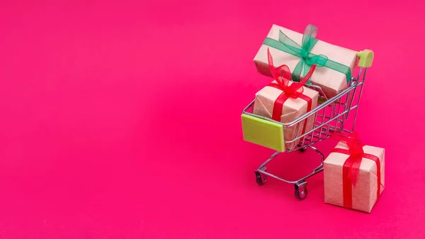 Small grocery cart with gift boxes on red-pink background. Gifts for Valentines day, Christmas and birthday. Shopping online. Holiday sales and discounts for New year. Retail and wholesale purchases.