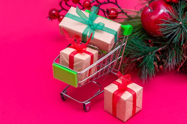 Petit chariot d'épicerie avec boîtes-cadeaux sur fond rouge-rose avec sapin de Noël. Shopping en ligne. Vente de vacances et réductions pour le Nouvel An. Achats au détail et en gros. Opérations commerciales. — Photo