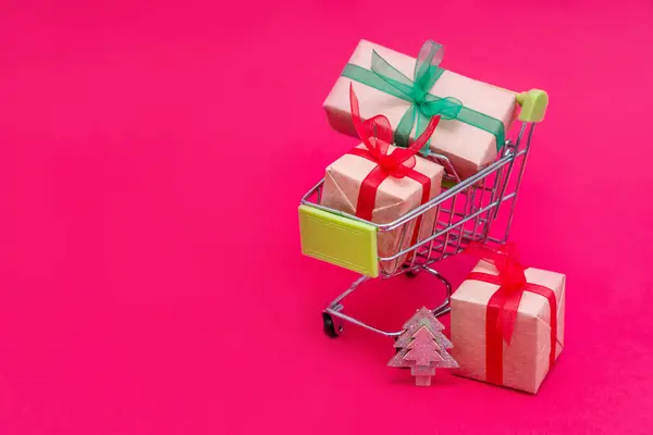 Petit chariot d'épicerie avec boîtes cadeaux de Noël sur fond rose. Shopping en ligne. Vente de vacances et réductions pour la nouvelle année. Achats au détail et en gros. Le commerce. Espace de copie. — Photo