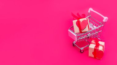Kırmızı-pembe arka planda hediye kutuları olan küçük bir market arabası. Sevgililer Günü 'nde, Noel' de ve doğum gününde sevgiyle hediye ver. İnternetten alışveriş. Tatil satışları ve indirimler. Perakende ve toptan alım.
