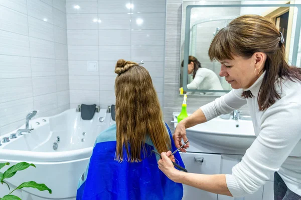 Home salon haircut during the quarantine. mature woman cuts long hair to young girl in bathroom. Mother is a hairdresser, cuts her daughters hair because of pandemic. Home pastime, leisure.