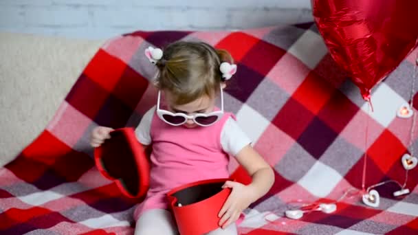 Mała piękna dziewczynka w okularach gra, otwiera czerwone pudełko w kształcie serca, wyciąga zabawkę i zamyka go. Uroczysta atmosfera. Walentynki. Miłość dziecka. Świętowanie urodzin. — Wideo stockowe
