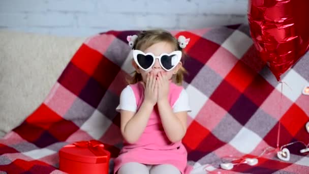 漂亮的小女孩戴着眼镜坐在沙发上 感到很惊讶 红色礼品盒 气球形 节日气氛 情人节假期 孩子的爱庆祝生日 — 图库视频影像