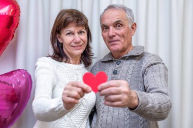 Mutlu çiftler Sevgililer Günü 'nü kutluyor. Erkek ve kadın kırmızı kalbi ellerinde tutar ve gülümserler. Romantik ilişkiler, aşk ve emeklilikte flört. Yaşlıların yaşam tarzı işte. Olgun emekliler.
