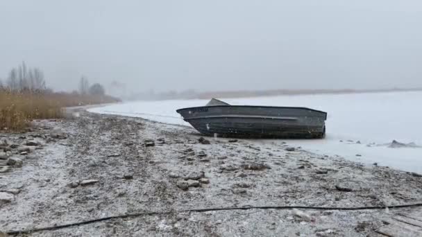 Pokryta śniegiem stara metalowa łódź rybacka leży w śniegu na brzegu zamarzniętego morza bez ludzi. Sezon połowowy dobiegł końca. Śnieg na jeziorze lub rzece. — Wideo stockowe
