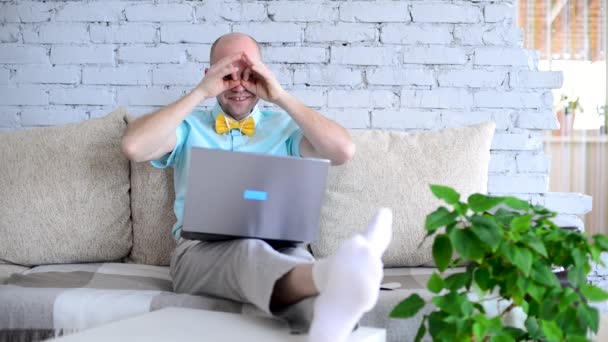 Crazy, cheerful, elegant man in shirt and bow tie fooling around and looking through binoculars made from his own hands on virtual date via call. Happy man talking on video chat in self-isolation. — Stock Video