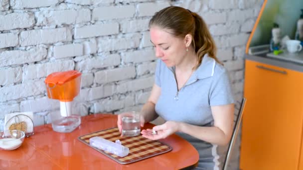 A jovem toma comprimidos. Sofrendo de mal-estar e dor, sentado à mesa da cozinha, lava o punhado de cápsulas com água. Tomar suplementos dietéticos e nutracêuticos. Curso do tratamento. — Vídeo de Stock