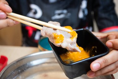 Shabu-Shabu restoranında ince dilimlenmiş et, bir tabağın üzerinde yemek çubuklarıyla tutulmuş. Arka planda sıcak bir tencere ve kaynar çorba var..