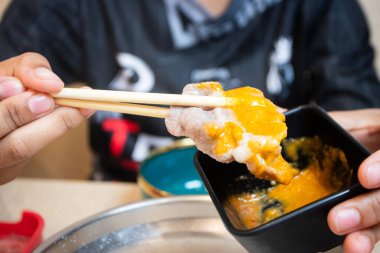 Shabu-Shabu restoranında ince dilimlenmiş et, bir tabağın üzerinde yemek çubuklarıyla tutulmuş. Arka planda sıcak bir tencere ve kaynar çorba var..