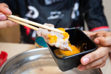 Shabu-Shabu restoranında ince dilimlenmiş et, bir tabağın üzerinde yemek çubuklarıyla tutulmuş. Arka planda sıcak bir tencere ve kaynar çorba var..