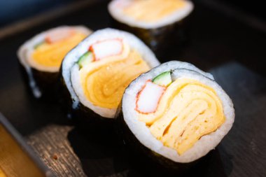 Yengeç çubuğu ve salatalıklı Japon Tamago yumurtalı suşi. Lezzetli maki suşisi, siyah bir tabakta servis edilir, içinde tatlı omlet ve pirinç bulunur..