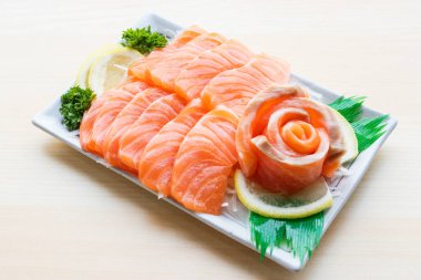 Beyaz tabakta limon ve maydanozla servis edilmiş taze somon balığı dilimleri. Parlak ahşap arka planda birinci sınıf Japon çiğ balık aperatifi..