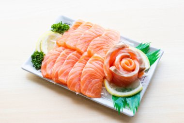 Beyaz tabakta limon ve maydanozla servis edilmiş taze somon balığı dilimleri. Parlak ahşap arka planda birinci sınıf Japon çiğ balık aperatifi..