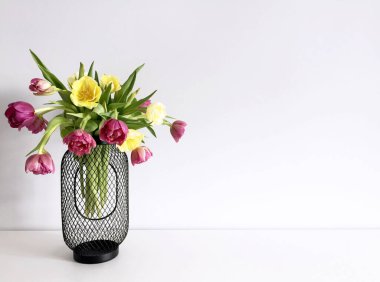 Beyaz bir masada gri bir duvara karşı siyah bir vazoda pembe ve sarı laleler.