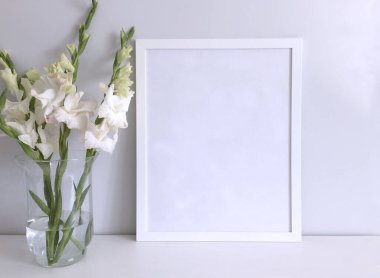 Çerçeve, yanında büyük cam bir vazo içinde beyaz bir gladioli buketi bulunan beyaz bir masa yüzeyindeki fotoğraf ya da resim için bir şablondur..