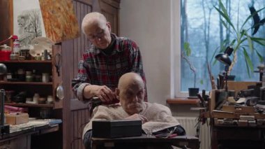 Yaşlı, gri saçlı, tekerlekli sandalyedeki yaşlı bir adam evinde tıraş makinesiyle karısının saçını kestiriyor.