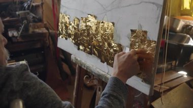 Tekerlekli sandalyedeki kıdemli sanatçı tuvaldeki yağlı boyaya altın sürüyor.