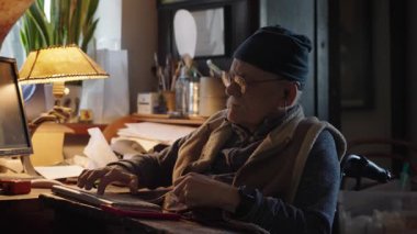 Kronik engelli yaşlı yetişkin tekerlekli sandalyede oturan bilgisayarla çalışıyor.