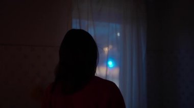 Genç bir kadın evinin penceresinden dışarı bakıyor. Odaklanamayan polis ışıklarıyla olay yerini inceliyor.