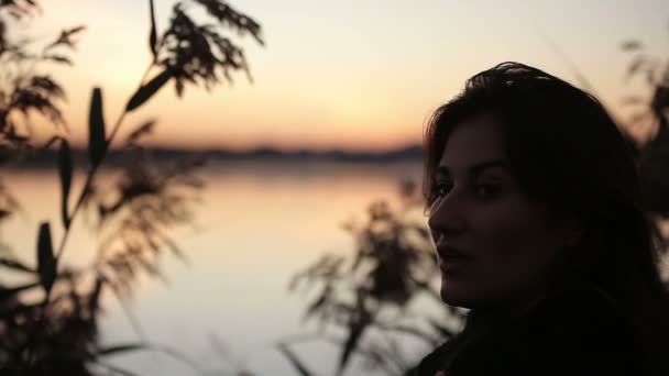 Mladá hezká brunetka s trávou sledovat východ slunce nad jezerem — Stock video