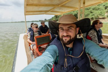 Şapkalı genç bir adam turistlerle teknenin selfie 'sini çekiyor. 14 Mart 2018 'de Kraliçe Elizabeth Ulusal Parkı, Uganda