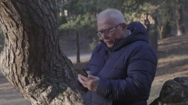 Gözlüklü neşeli emekli telefonda ağaç gövdesiyle konuşuyor.
