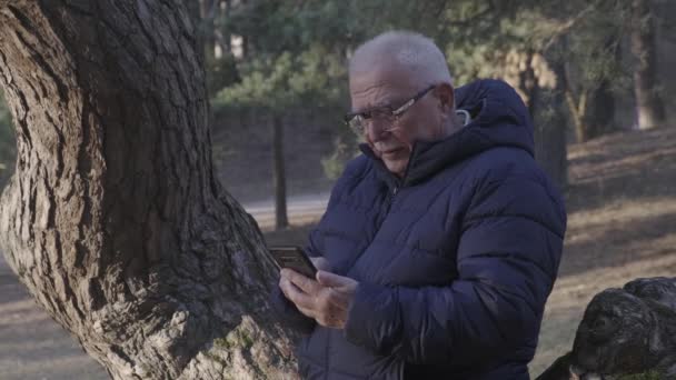 Veselý důchodce v brýlích mluví na telefonu po kmeni stromu — Stock video