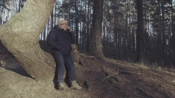 穿着夹克和眼镜的退休人员靠在树上讲电话 — 图库视频影像