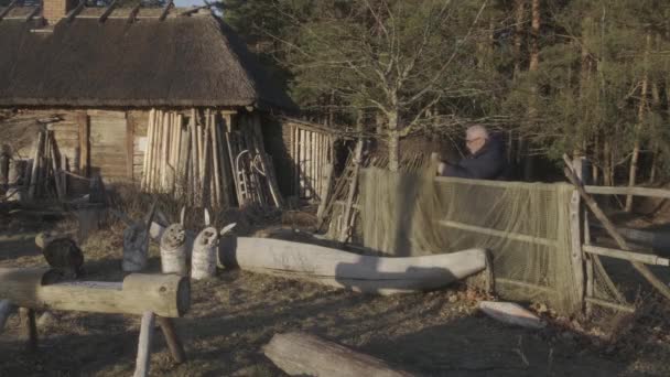 Человек распутывает рыболовные сети на заборе против старой деревянной хижины — стоковое видео