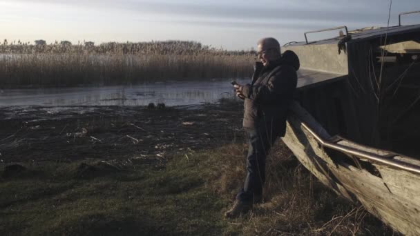 Pensionär upplyst av solen står vid gammal båt och talar i telefon — Stockvideo