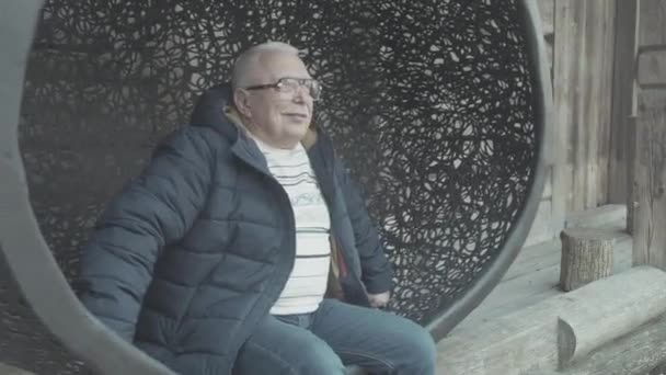 Senior man in winterjas zit in opknoping cocon stoel — Stockvideo