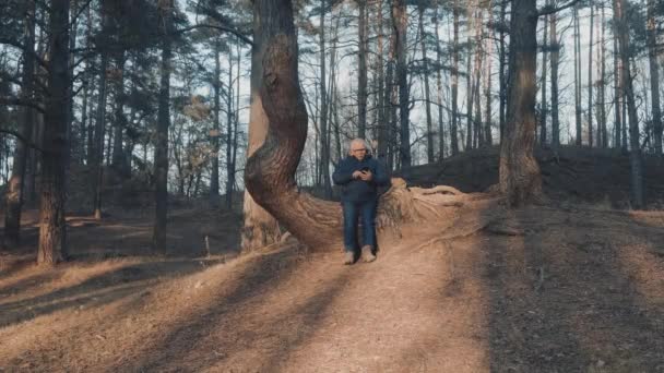 Elderly man sits on crooked tree dials number and calls — Stock Video